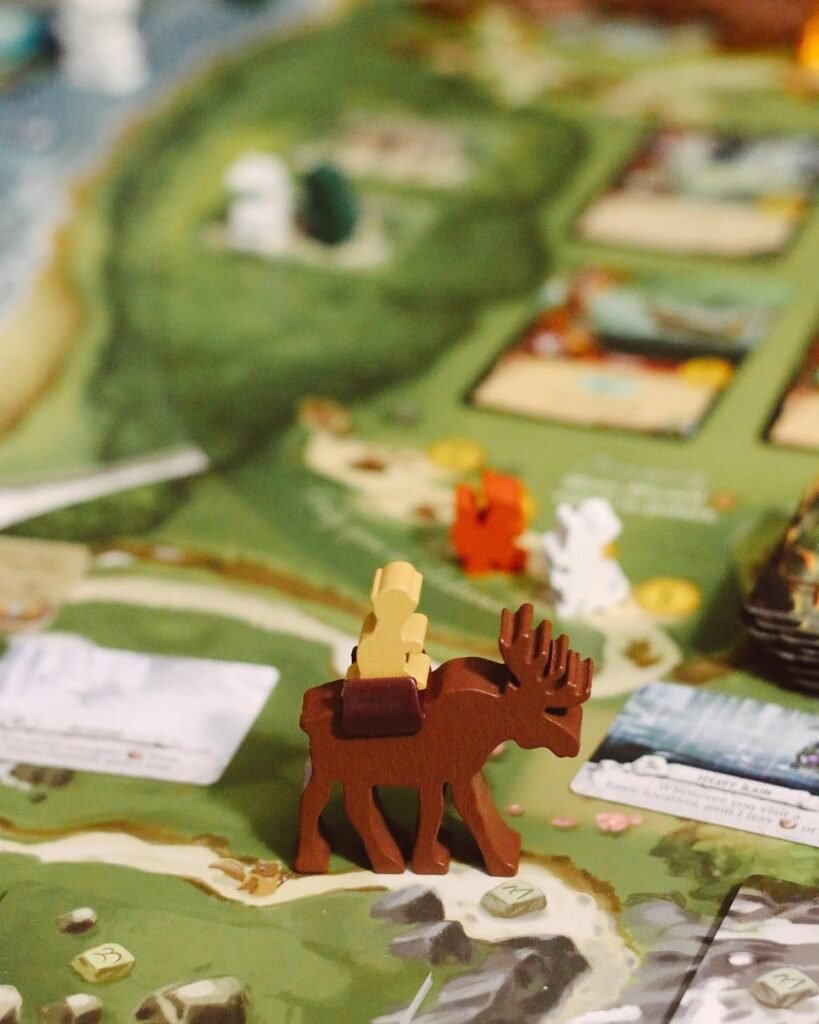 Prim-plan cu tabla Everdell: o figurină de lemn în formă de elan maro poartă un meeple muncitor deschis la culoare peste un peisaj de pădure verde, printre jetoane de resurse și cărți.