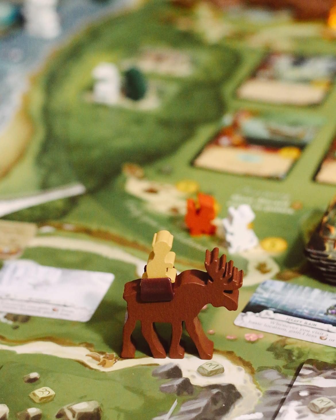 Prim-plan cu tabla Everdell: o figurină de lemn în formă de elan maro poartă un meeple muncitor deschis la culoare peste un peisaj de pădure verde, printre jetoane de resurse și cărți.