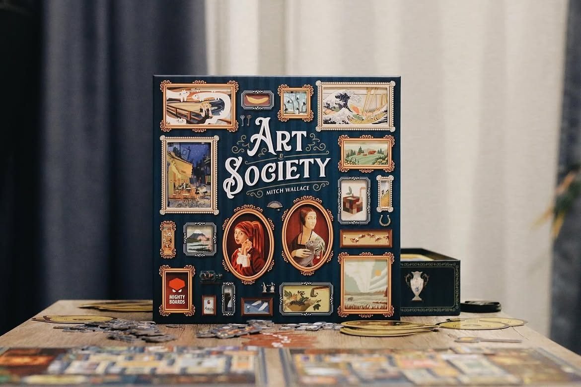 Cutia jocului de societate „Art Society” așezată vertical pe o masă de lemn, cu coperta plină de tablouri înrămate în miniatură și jetoane de carton împrăștiate în față.