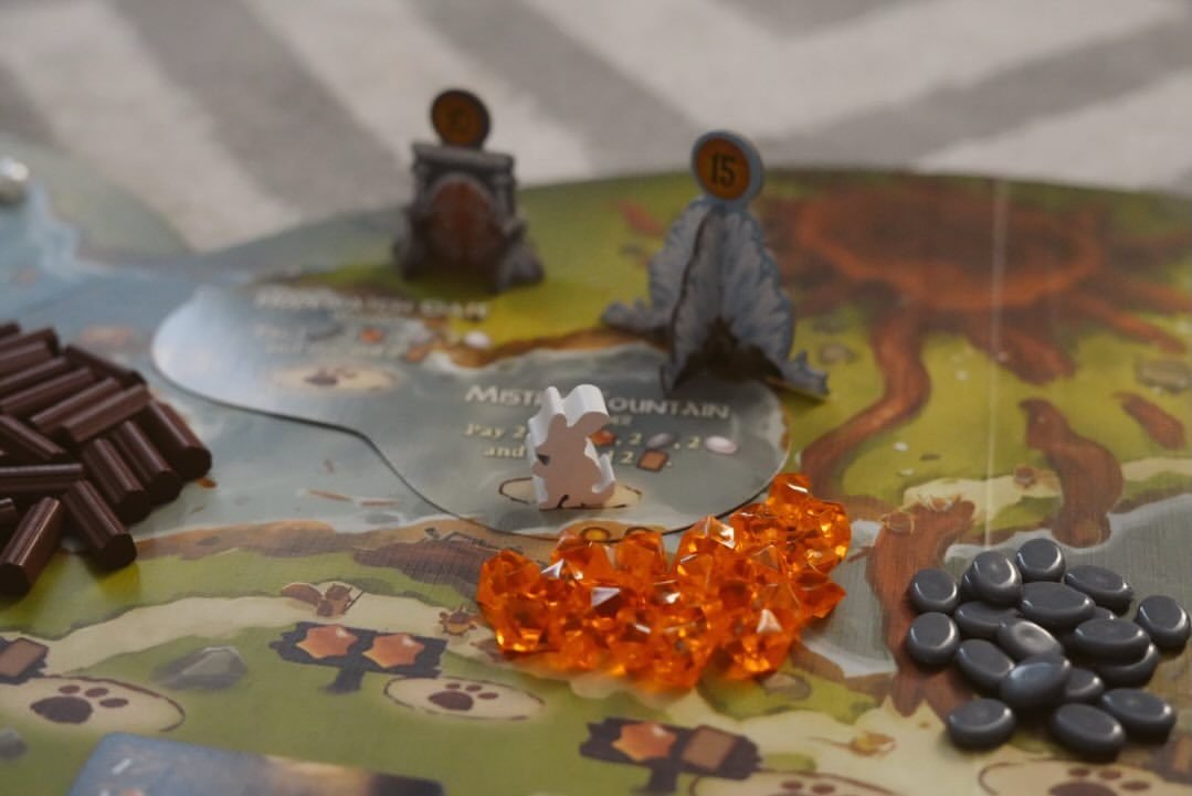 Vedere macro a componentelor Everdell: un meeple muncitor iepure alb lângă standuri de munte din carton, cu grămezi de jetoane de resurse — lemn maro, cristale portocalii și pietre gri.