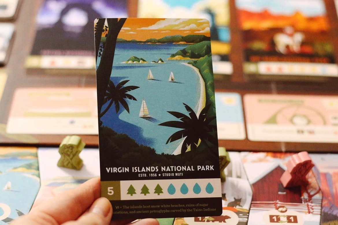 O mână ține cartea „Virgin Islands National Park” din Parks, un golf stilizat cu bărci cu pânze și palmieri, deasupra tablei de joc neclare.