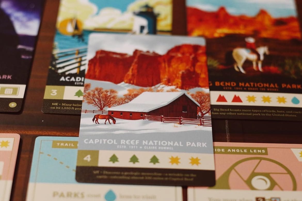 Cartea „Capitol Reef National Park” din Parks, cu un hambar roșu și un cal în zăpadă sub stânci roșii, așezată peste alte cărți de parcuri naționale.