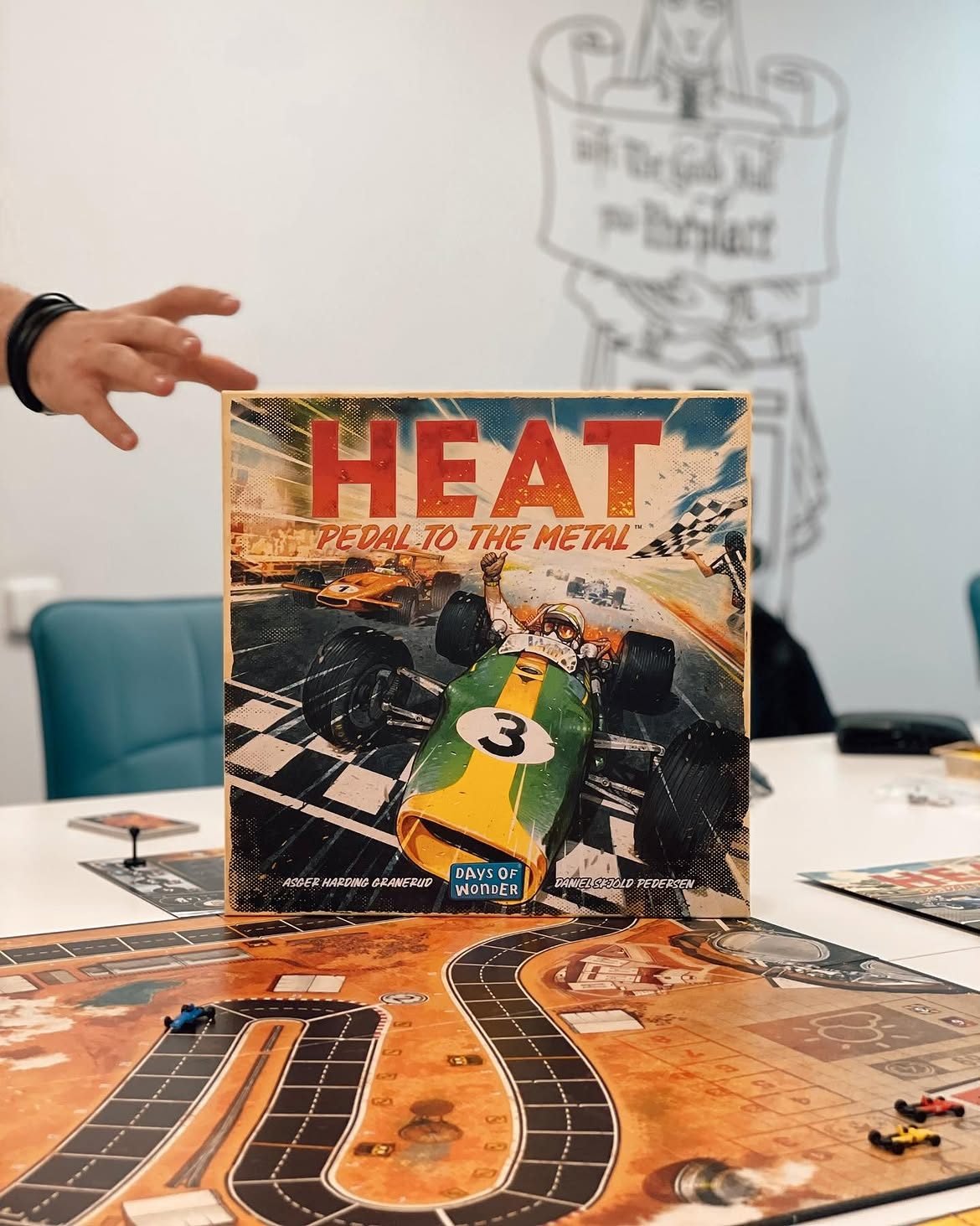 Cutia „Heat: Pedal to the Metal” stă pe tabla-circuit de curse, în timp ce o mână intră în cadru din lateral; pe perete atârnă un banner desenat în linii.