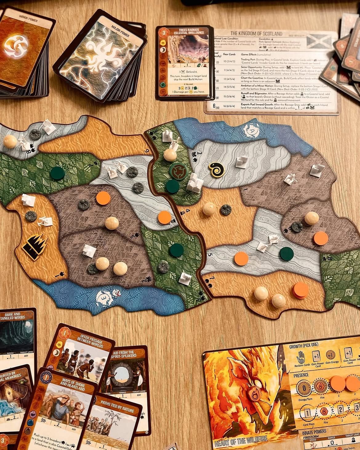 Vedere de sus a unei partide Spirit Island: o tablă-insulă din două panouri cu piese de lemn și jetoane de invadatori, înconjurată de cărți de putere, o fișă de adversar și un panou de spirit al focului.
