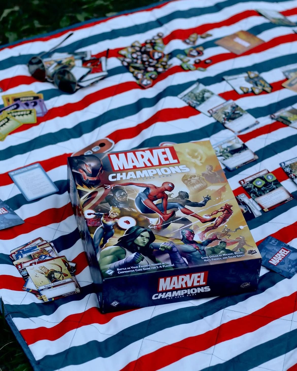 Cutia „Marvel Champions: The Card Game” pe o pătură de picnic în dungi roșii și albe, în aer liber, înconjurată de cărți de eroi și inamici, jetoane și ochelari de soare.